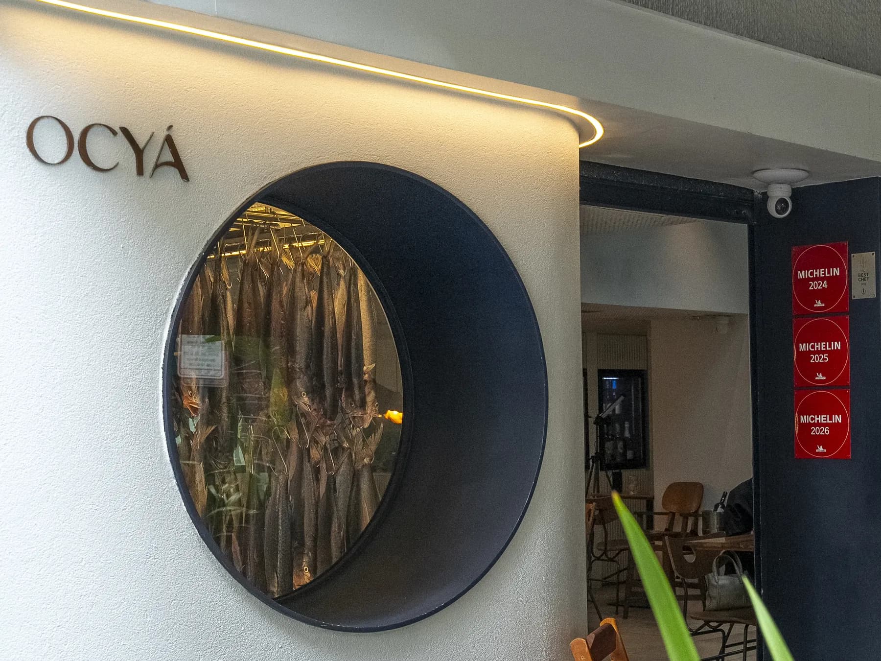 Fachada do Ocyá Leblon: a logo, a câmara de maturação com peixes pendurados e, na entrada, as placas do Guia MICHELIN de 2024, 2025 e 2026